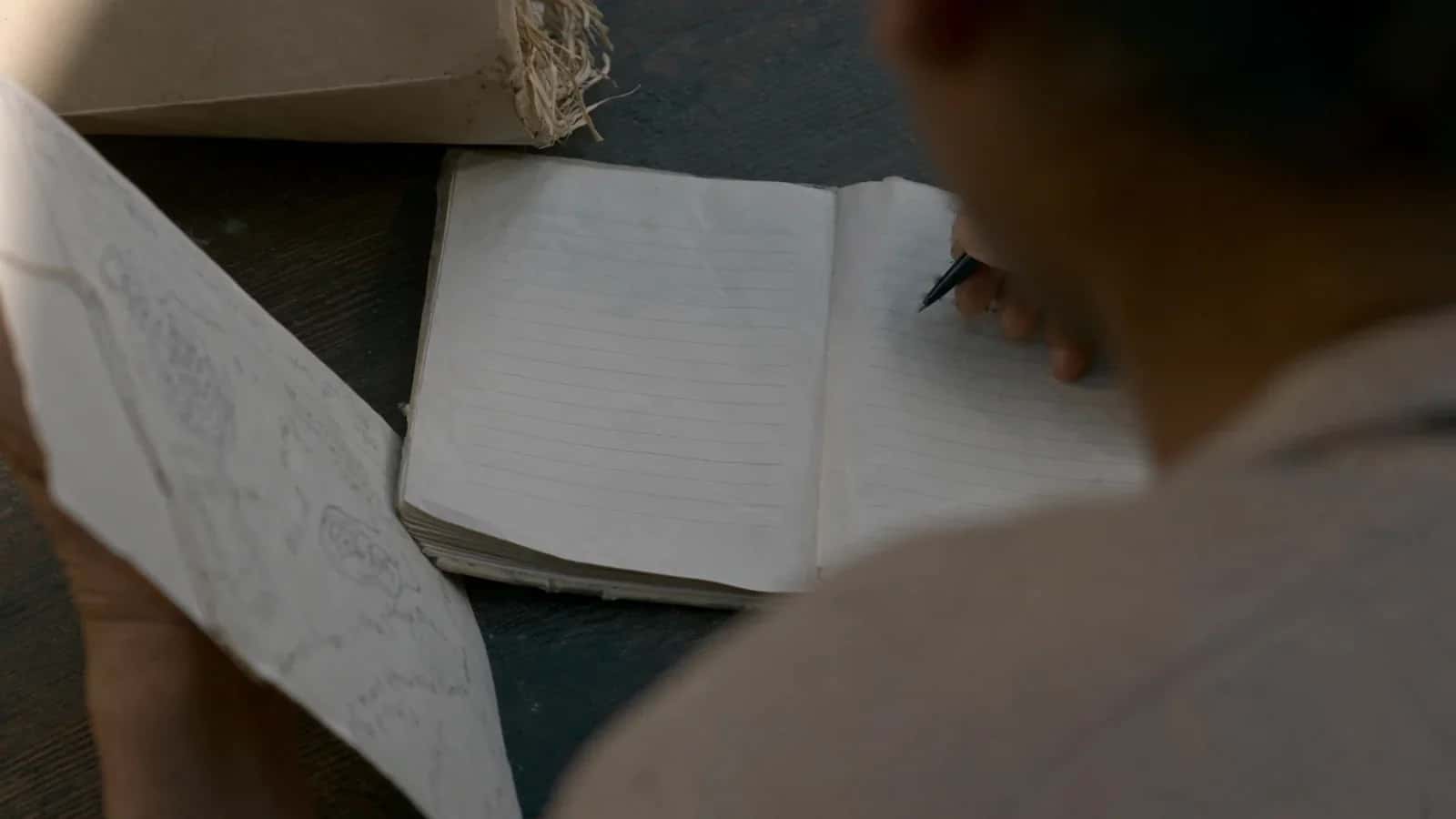 Una persona empieza a escribir en una libreta abierta; a su lado, otra sostiene un papel con bocetos a lápiz