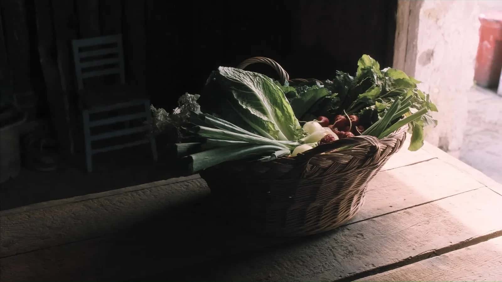 Cesta de mimbre con verduras de hoja y rábanos sobre una mesa, junto a una ventana